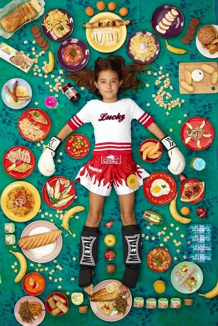 Rosalie en tenue de boxe, entourée de pain, de fruits et d’assiettes sur un fond turquoise.